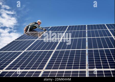 Instantané horizontal de l'ouvrier penché en uniforme et casque, debout sur l'échelle sur le dessus de la plante solaire, l'installation de modules solaires le jour ensoleillé. Concept de sources d'énergie alternatives et innovations. Banque D'Images