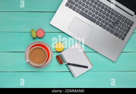 Ordinateur portable, macarons colorés, biscuits, tasse de café, carnet avec un stylo sur une table en bois bleu. Vue de dessus. Pose à plat Banque D'Images