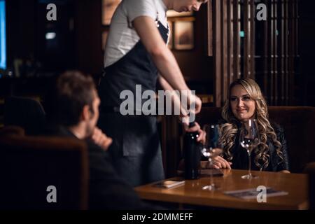 le barman verse du vin rouge d'une bouteille à une femme et à un homme assis à une table. Banque D'Images