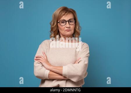Femme caucasienne âgée sérieuse en lunettes avec les mains pliées. Studio Headshot isolé sur fond bleu. Banque D'Images
