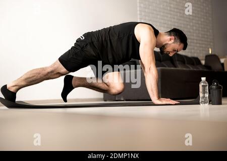 Homme faisant des exercices d'alpiniste sur tapis de yoga noir. Entraînement du matin. Salon moderne blanc sur fond. Bouteille d'eau en plastique. Entraînement intensif Banque D'Images