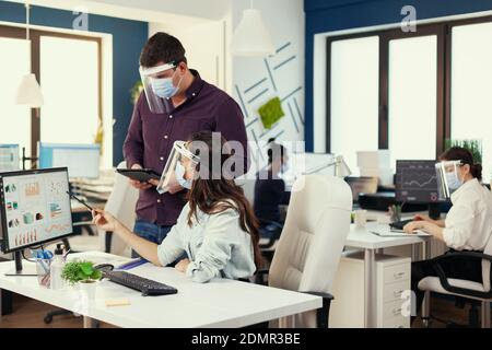 Company analysts helping each other to finish a project at workplace wearing face mask as safety precaution for covid19. Multiethnic team in new normal financial office in corporate company typing on computer, taking notes on tablet. Stock Photo