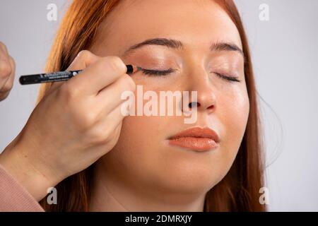 Gros plan photo de maquillage pour une fille aux cheveux rouges, yeux fermés et un crayon cosmétique pour les yeux. Concept de beauté. Banque D'Images