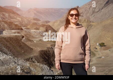 Bonne femme brune avec des lunettes de soleil élégantes dans le sweat à capuche regarde appareil photo et bénéficie d'un paysage merveilleux de vallée vallonnée avec la route en haute montagne le jour ensoleillé Banque D'Images