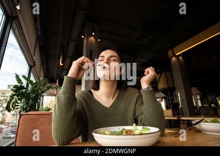 Une jeune femme satisfaite aime la salade savoureuse dans un restaurant, en mettant de la fourchette dans sa bouche et en fermant les yeux. Bon service client. Une alimentation saine concep Banque D'Images