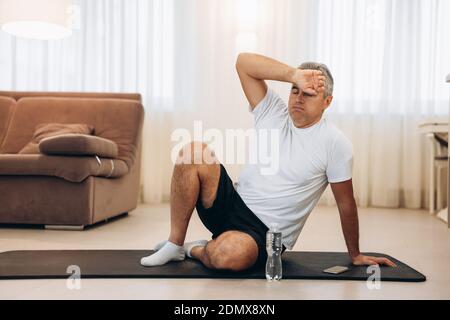 L'homme âgé essuye la sueur de son front après un entraînement à la maison difficile. Concept de mode de vie sain. Séjour en arrière-plan. Homme fatigué après un entraînement intensif Banque D'Images