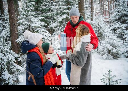 Un beau jeune couple avec des enfants marche à travers une fabuleuse forêt d'hiver pendant le nouvel an ou les vacances de Noël. Ils sont réchauffés par du thé chaud dans des thermos Banque D'Images