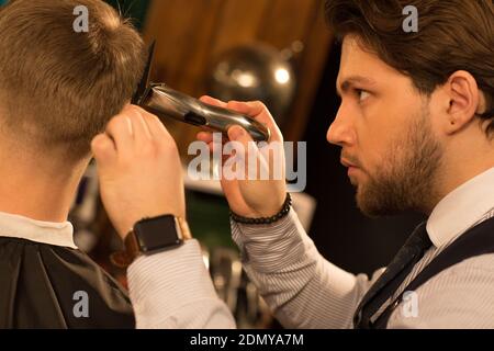 Photo courte en gros plan d'un jeune professionnel barber a l'air concentré donnant une coupe de cheveux à son client utilisant tondeuse clipper equi Banque D'Images