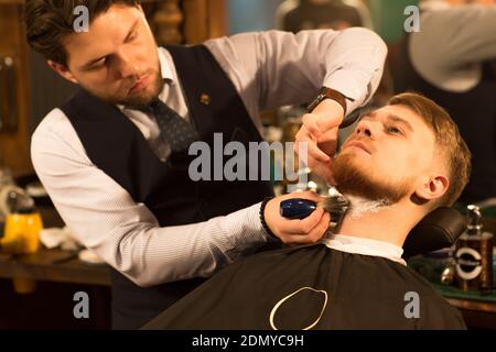 Beau jeune barbu rasé par un barbier professionnel au barbershop appliquant de la crème de rasage sur le cou de sa profession de client servi Banque D'Images