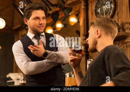 Beau coiffeur professionnel joyeux parlant à son client après avoir donné il a une nouvelle coupe de cheveux travaillant à la mode de style barbershop service hippster mas Banque D'Images