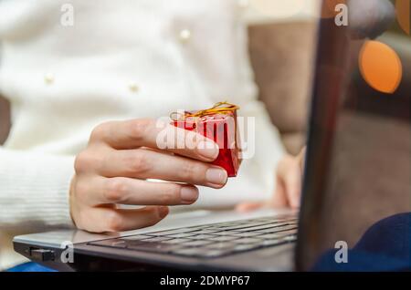 Une femme dans un chandail blanc tient un ordinateur portable sur ses genoux et tient une boîte cadeau rouge dans sa main en gros plan. Concept de Noël, nouvel an, vacances Banque D'Images