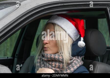 Jeune femme blonde dans un chapeau de Père Noël est assis au volant d'une voiture et regarde avec un regard sérieux et triste dans la distance Banque D'Images