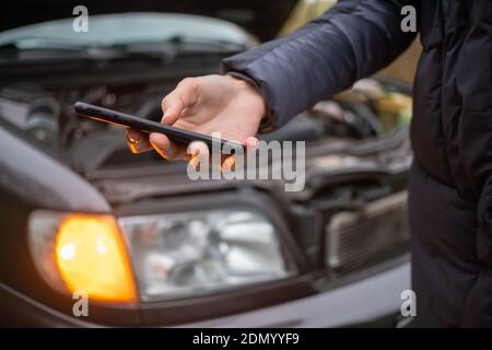 La main d'une femme dans une veste peut contenir un téléphone portable noir gros plan contre le capot ouvert d'une voiture sur un arrêt d'urgence Banque D'Images