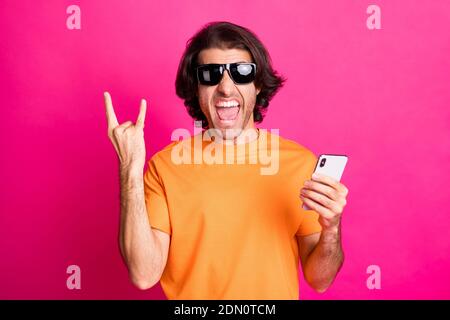 Photo d'un jeune homme fou tenir le téléphone bouche ouverte spectacle les cornes portent un t-shirt orange lunettes de soleil isolé de couleur rose fond Banque D'Images