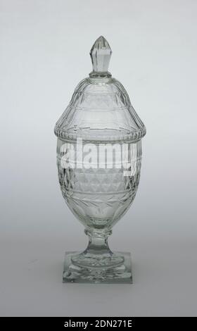 Jar and lid, Glass, Tall urn-shaped body; tapered, fluted stem; square base with star cutting on bottom; tall domed cover with faceted finial; body cut with row of narrow flutes, band of diamonds, a garland circlet, wide flutes down to base; cover cut with conforming pattern; glass greenish and bubly, heavy., Waterford, Ireland, ca. 1800, glasswares, Decorative Arts, Jar and lid Stock Photo