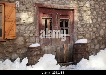 Plan horizontal de la neige devant la porte en bois abandonnée d’une maison de montagne flanquée de deux barils couverts de neige, Eskunhau, Vall d’Aran Banque D'Images