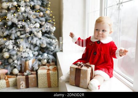 Une petite fille à tête rouge dans un costume de Noël dans une robe rouge se trouve sur le rebord de la fenêtre près de l'arbre de noël avec boîte cadeau de noël. Banque D'Images