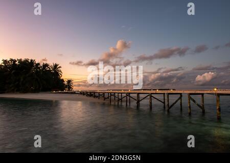 Jetée en bois avec des lanternes sur l'océan s'étendant de l'île tropicale, avec l'eau floue et le coucher du soleil en arrière-plan, Maldives, longue exposition. Banque D'Images