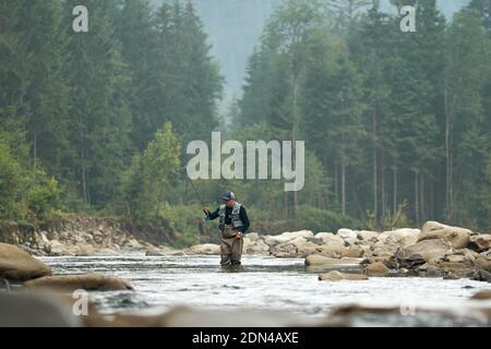 Pêcheur dans des vêtements spéciaux et des bottes utilisant la tige professionnelle pour attraper le poisson à la rivière. Homme debout dans l'eau parmi les montagnes vertes. Con. Relaxation Banque D'Images