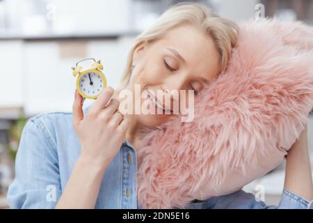 Gros plan d'une belle femme heureuse tenant un petit réveil, allongé sur un oreiller rose moelleux. Charmante femme prenant une petite sieste Banque D'Images