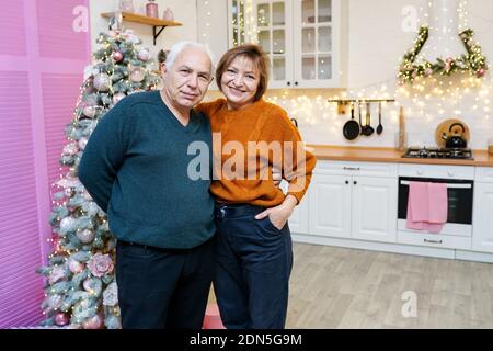 Vieux heureux couple à la maison sur la cuisine blanche avec arbre de Noël et guirlandes décorées roses. Banque D'Images