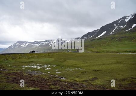 Une voiture se déplace le long d'une section de route surélevée de la ville de Seydisfjordur en Islande, par une journée de dépassement. Des montagnes enneigées bordent la route. Banque D'Images