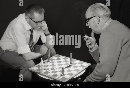 1960s, historique, deux hommes plus âgés se concentrant comme ils s'assoient en face l'un de l'autre jouant un jeu d'échecs, avec le chessboard assis sur une petite table de café les brochettes, avec seulement quelques pièces restantes sur le plateau, Angleterre, Royaume-Uni. Un jeu de société populaire, un jeu de stratégie abstrait, la version moderne datant du 15th siècle, le jeu d'échecs est joué sur un chessboard de 64 carrés avec 16 pièces, le vainqueur attaque et costère le roi de l'adversaire, donc il n'y a aucun moyen sur le plateau pour qu'il s'échappe. Banque D'Images