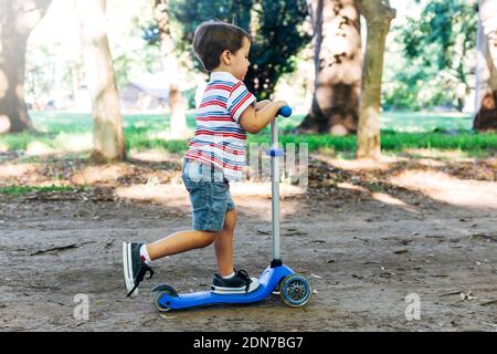 Enfant sur un scooter dans le parc. Les enfants apprennent à skate roller board. Petit garçon patinant le jour ensoleillé de l'été. Activités de plein air pour les enfants dans une rue sûre. Banque D'Images