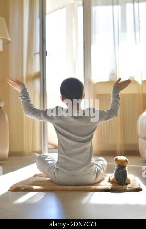 L'enfant s'assoit avec son dos à la maison. Sport de fitness yoga sur le tapis avec peluche chien. Méditation dans la position du lotus avec les bras ouverts, sur le sol Banque D'Images