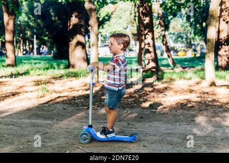 Enfant sur un scooter dans le parc. Les enfants apprennent à skate roller board. Petit garçon patinant le jour ensoleillé de l'été. Activités de plein air pour les enfants dans une rue sûre. Banque D'Images