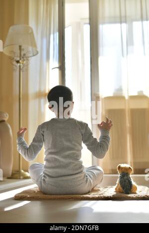 L'enfant s'assoit avec son dos à la maison. Entraînement physique yoga sur le tapis avec peluche chien. La méditation dans la position du lotus avec les mains fait mudra, sur le floo Banque D'Images