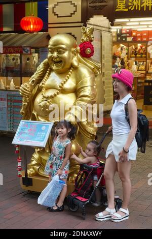 A l'extérieur d'une boutique d'objets rituels bouddhistes à Waterloo St., Singapour, une femme et des filles posent à une statue du Bouddha riant (Budai / Hotei) Banque D'Images