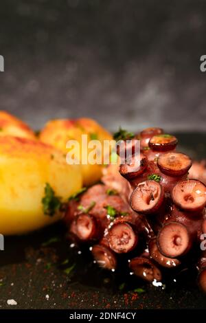 Grilled octopus tentacles with potatoes seasoned with Spanish paprika, olive oil, parsley and sea salt on a black slab and fancy background. Gourmet k Stock Photo