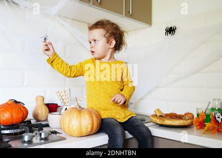 Cute little girl sitting on kitchen table and holding halloween dacorations spider Stock Photo