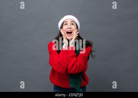 Excitée belle femme asiatique en rouge chandail de Noël et chapeau crier avec les mains entourant la bouche dans un gris isolé en studio arrière-plan Banque D'Images