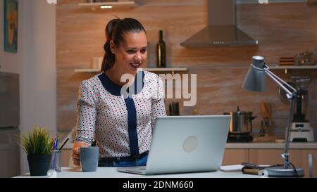 Femme entrepreneur à distance ayant une vidéoconférence avec des collègues utilisant un ordinateur portable assis dans la cuisine tard dans la nuit. Employé occupé utilisant un réseau sans fil moderne faisant des heures supplémentaires pour le travail. Banque D'Images