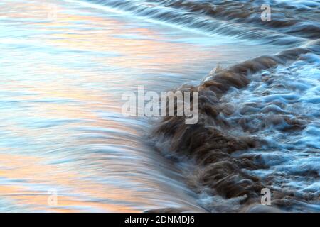 La marée se déverse sur les échalotes d'une plage au crépuscule, reflétant les couleurs du coucher de soleil dans l'eau en mouvement rapide. Banque D'Images