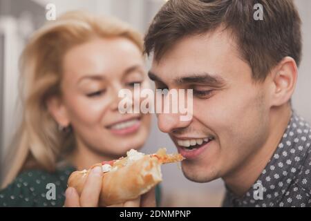Gros plan d'un jeune homme joyeux et charmant piquant une pizza, sa belle petite amie tient. Couple heureux appréciant manger à la maison, en commandant p Banque D'Images