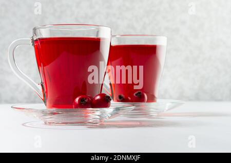 Thé aux herbes rouge d'automne dans deux tasses transparentes avec baies d'aubépine mûres au soleil sur table en bois blanc, gros plan. Banque D'Images