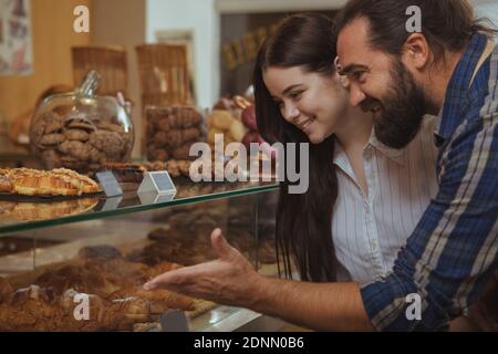 Photo d'un joli boulanger à barbes et joyeux, en train de travailler dans son café, aidant ainsi une cliente. Belle femme heureuse souriant, choisissant le passé Banque D'Images