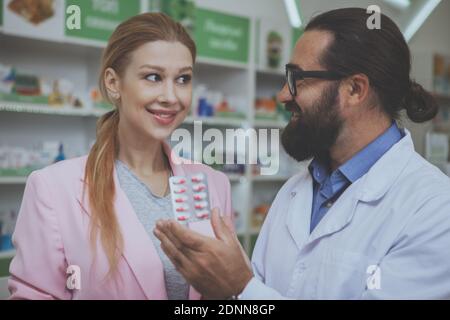 Belle jeune femme souriant, achetant des pilules à la pharmacie. Pharmacien mature barbu aidant sa cliente féminine à montrer sa plaquette de pilules. Femme Banque D'Images