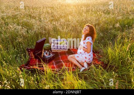 Une femme enceinte est assise sur la couverture rouge près d'un gramophone et d'un panier de fruits sur le champ de blé par temps ensoleillé. Banque D'Images