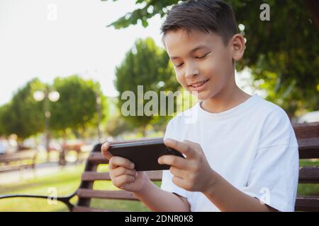 Charmant jeune garçon asiatique souriant, jouant en ligne sur son smartphone à l'extérieur, espace de copie. Un jeune garçon adorable qui apprécie le téléphone portable Banque D'Images