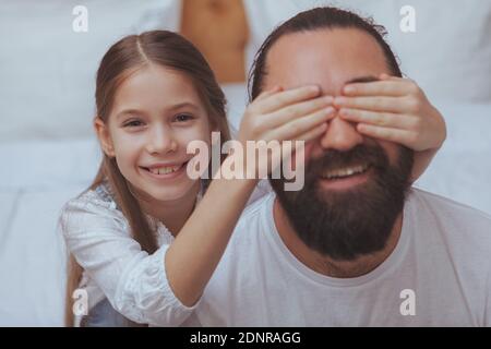 Gros plan d'un homme mûr barbu souriant, sa fille couvrant ses yeux avec ses mains. Joyeux homme mature surpris par sa petite fille. C Banque D'Images