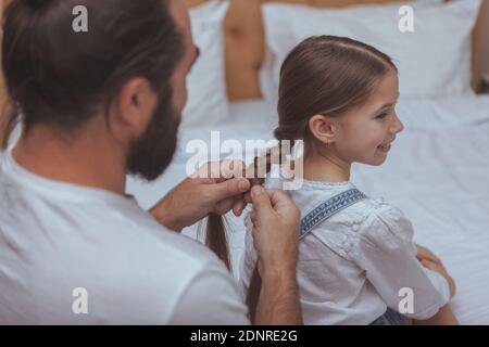 Vue arrière photo courte d'un homme barbu qui apprécie les cheveux tressés de sa belle fille. Adorable petite fille souriant, lui faisant passer ses cheveux dans des tresses Banque D'Images