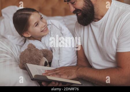 Adorable petite fille heureuse tenant un ours en peluche, souriant à son père, pendant qu'il lit son conte de fées Bonne nuit. Photo courte d'une main gaie Banque D'Images