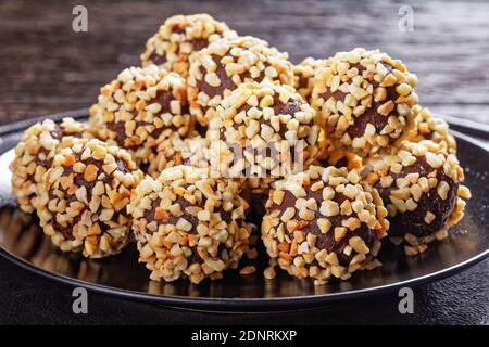 Truffes au chocolat noir enrobées d'amandes écrasées servies sur une assiette noire sur fond de bois foncé, vue de dessus, gros plan Banque D'Images