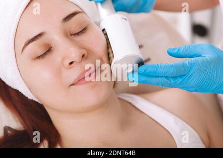 Un petit gros plan d'une jeune femme détendue souriant tout en obtenant un soin du visage resserré de la peau par une esthéticienne. Concept de thérapie Endosphères Banque D'Images