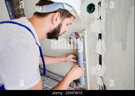 Gros plan du jeune barbu dans un casque de sécurité vissant la bride sur le tuyau de chauffage avec un tournevis. Homme installant un système d'eau dans l'appartement. Concentrez-vous. Travaux de plomberie, concept de rénovation de la maison Banque D'Images