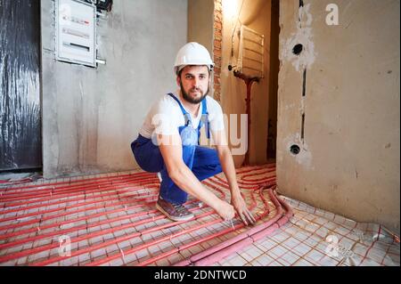 Homme travaillant dans un casque de sécurité en train de s'accroupir tout en installant des tuyaux pour le sol chaud dans l'appartement. Homme barbu plombier dans les combinaisons de travail installation du système de chauffage par le sol. Concept de rénovation de la maison. Banque D'Images